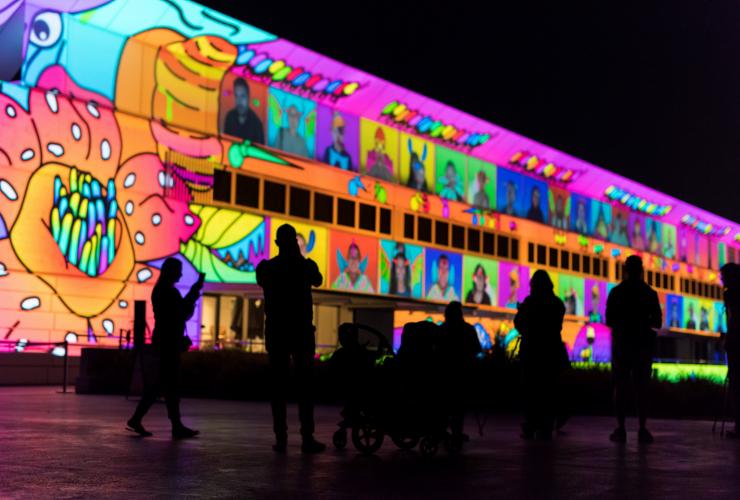 オーストラリア首都特別地域、キャンベラ、エンライテン・フェスティバルにて、建物の前面にカラフルなアートが浮かび上がるインスタレーション「イルミネーションズ」を前に立つ人々の黒いシルエット © Enlighten Festival
